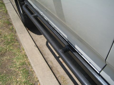 1990 Ford Bronco - black tubular side step bar mounted along the rocker of a light-colored vehicle, viewed over the curb and grass.
