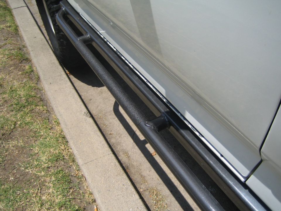 Black tubular side step bar mounted along the rocker panel of a vehicle by a curb