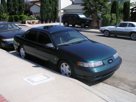 1993 Ford Taurus - A dark green 1993 Ford Taurus sedan is parked curbside on a sunny day, with other vehicles visible in the background.