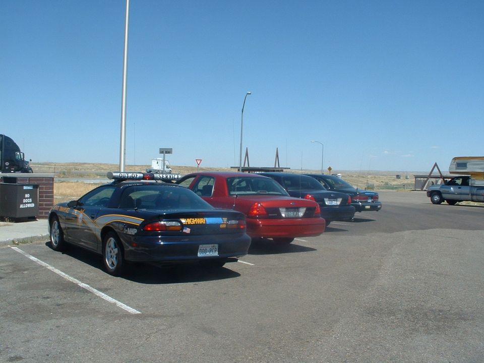 Four police cars parked in a lot, including a red Ford Crown Victoria and a dark blue Chevrolet Camaro with "HIGHWAY" markings