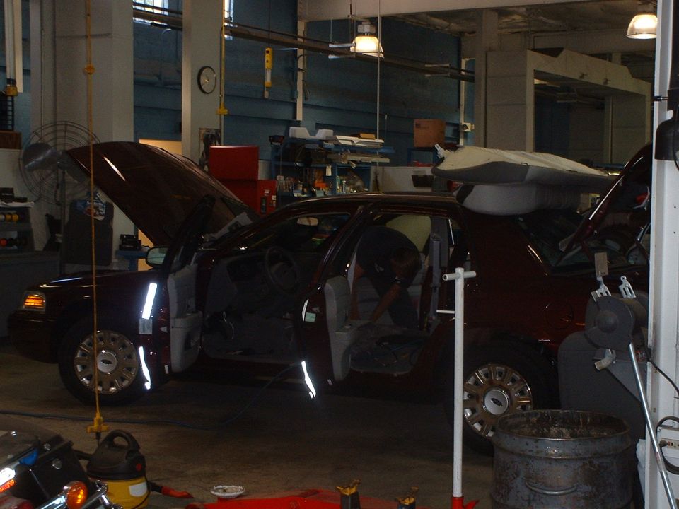 Dark sedan with doors and hood open inside a busy repair garage, person working inside