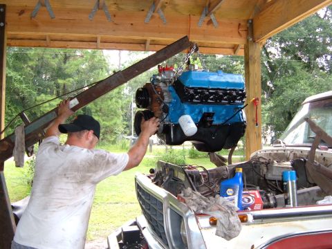 1978 Ford Bronco - A blue engine is being carefully lowered into the engine bay of a truck using an engine hoist.
