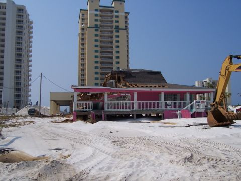 1995 Ford Bronco - An excavator sits near a damaged beach house, with tall buildings visible behind it under a clear blue sky.