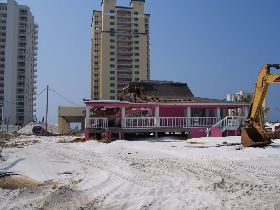 damaged pink beach house with excavator nearby, tall buildings in background