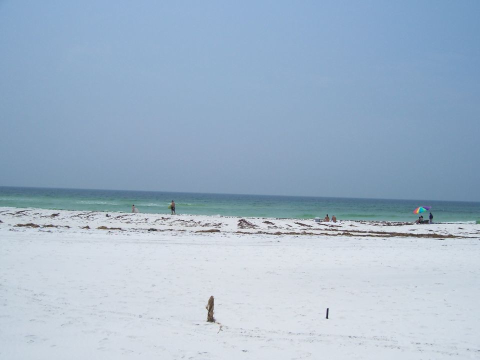 Beach with white sand, turquoise water, and a clear blue sky. People are visible on the shore and in the water