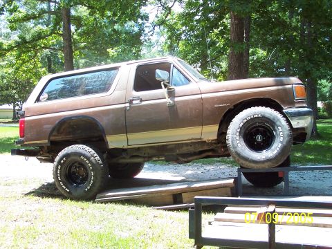 1987 Ford Bronco - Side profile of a brown and tan two-tone full-size Ford Bronco with removable hardtop, parked on wooden planks over gravel in a shaded yard…