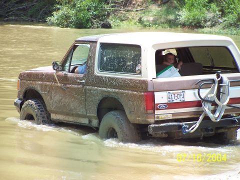 1987 Ford Bronco - A brown 1987 Ford Bronco navigates through a muddy waterway, with a child visible in the back seat.