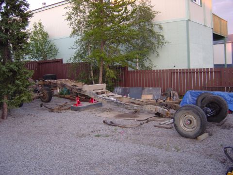 1986 Ford F-150 - The chassis of a 1986 Ford F-150 is visible, disassembled and resting on gravel with a building and trees in the background.