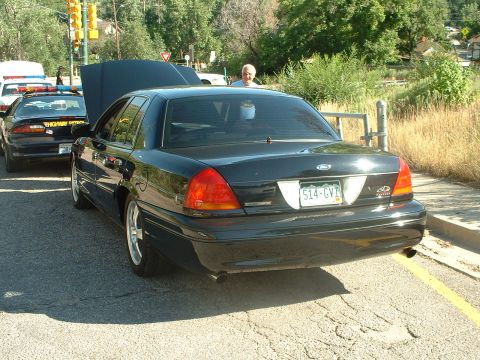 1999 Ford Crown Victoria - A black sedan with its hood open is parked next to a highway patrol car.