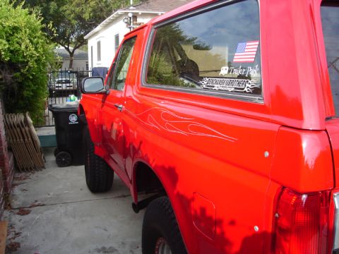 1990 Ford Bronco - Side view of a lifted red Ford Bronco SUV with silver flame pinstriping and window decals parked in a narrow driveway by a trash bin.