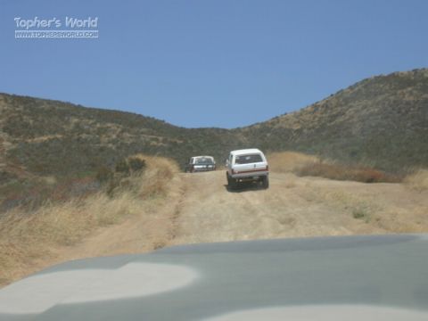 1990 Ford Bronco - Two light-colored SUVs head up a dusty dirt trail winding through dry, brush-covered hills under a clear blue sky.