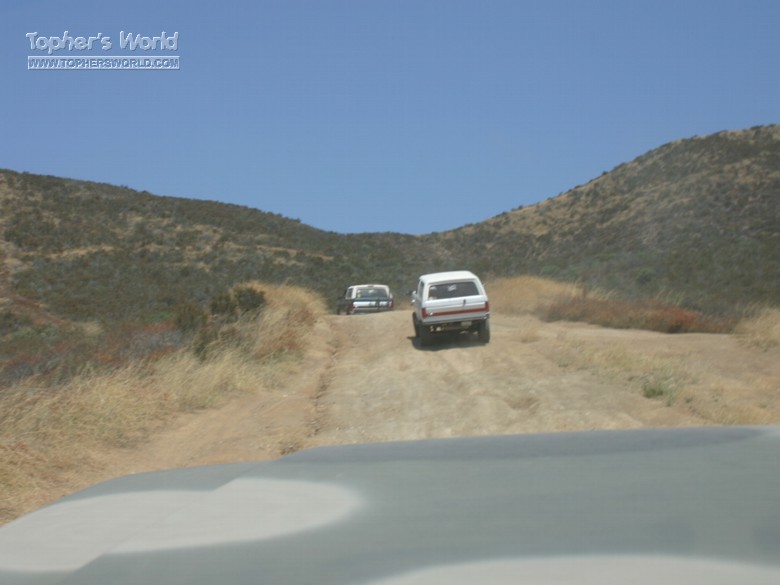 Two SUVs climbing a dirt trail through dry brushy hills, seen over a vehicle hood
