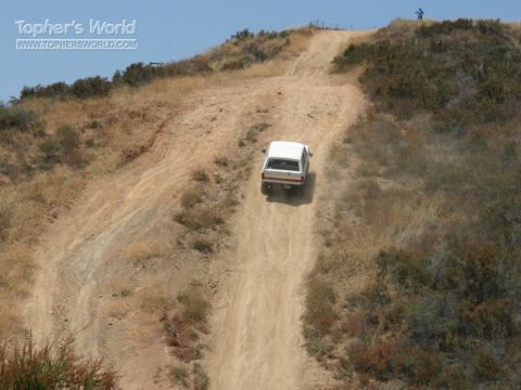 1990 Ford Bronco - white SUV makes its way down a steep, eroded dirt hillside trail amid dry scrub, with a tiny figure visible at the ridge.