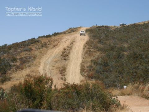 1990 Ford Bronco - small light-colored SUV climbs a steep sandy trail up a dry brush-covered hill, with another vehicle parked near the crest.