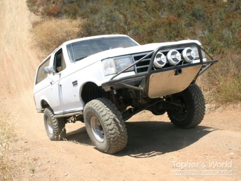 1990 Ford Bronco - lifted white Ford Bronco with a tubular front bumper, three round driving lights, and large mud tires sits on a dirt slope.