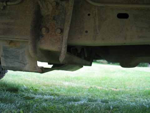 1995 Ford Bronco - Underbody shot taken from ground level looking beneath a truck parked on grass, showing a surface-rusted boxed frame rail, a bracket secure…