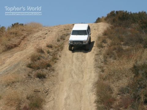 1990 Ford Bronco - lifted white SUV with a front brush guard descends a steep, rutted dirt hill against a clear blue sky.