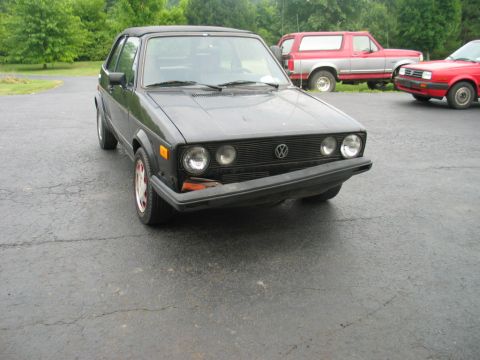1986 Volkswagen Cabriolet - Front three-quarter view of a black convertible, possibly a 1986 Volkswagen Cabriolet, parked on asphalt with other vehicles in the backgro…