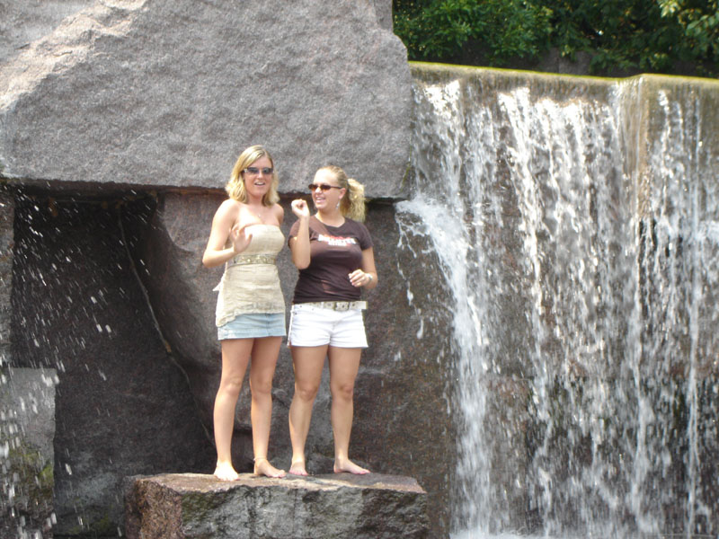 Two women stand on rocks near a waterfall. Water cascades down a stone wall