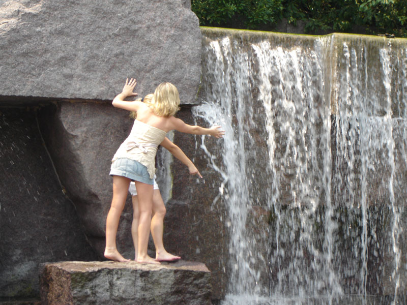 Two women stand on rocks near a waterfall, reaching out towards the water