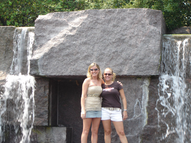 Two women stand in front of a large rock with waterfalls on either side. One wears a strapless top and denim skirt, the other a t-shirt and white shorts