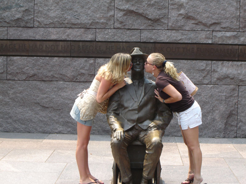 Statue of a seated man with two women kissing him in front of a stone wall with engraved text