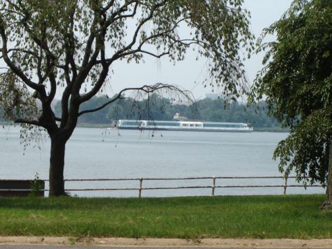 1986 Ford Bronco - A long white boat travels on the water, framed by trees and a grassy embankment.