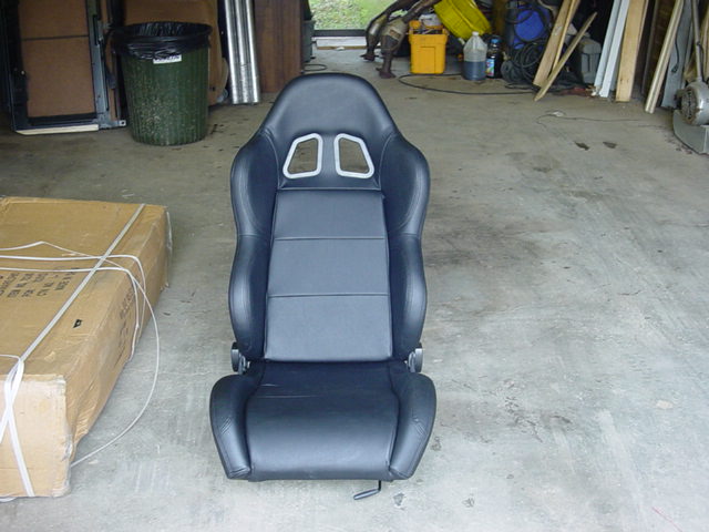Black racing car seat with silver trim around headrest openings, on concrete floor