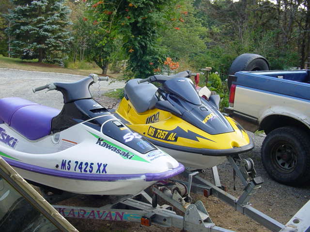 Two jet skis on a trailer, one white and purple, one yellow and blue, with a pickup truck behind them