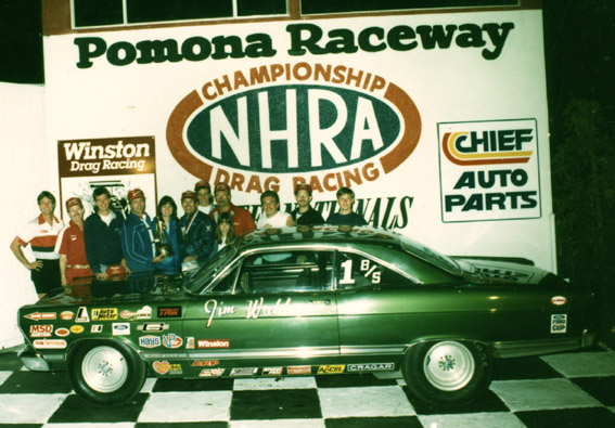 Dark green drag racing coupe with sponsor decals in NHRA Pomona Raceway winner's circle