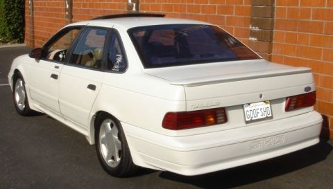 1991 Ford Taurus - White first-generation Ford Taurus SHO sedan viewed from the rear three-quarter beside a red brick wall, with a moonroof, decklid spoiler,…