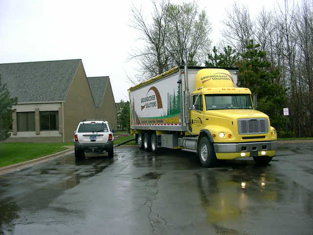 White SUV from rear, parked next to a yellow semi-truck with "GROUNDCOVER SOLUTIONS" text on its trailer