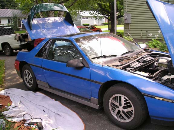 Blue 1984 Pontiac Fiero with hood and trunk open, parked on asphalt driveway