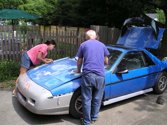 Two people sanding and prepping a blue Pontiac Fiero with raised engine hatch