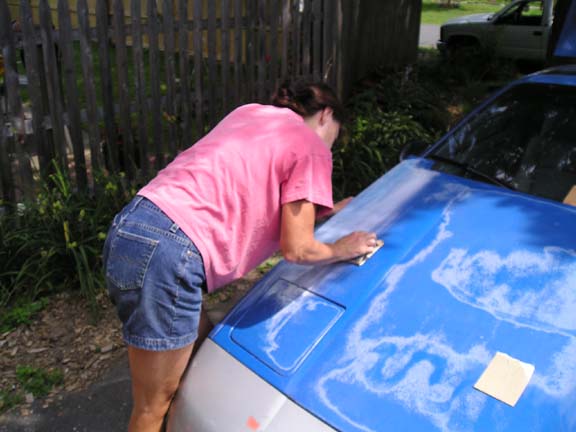 Person in pink shirt and denim shorts sanding the blue hood of a car
