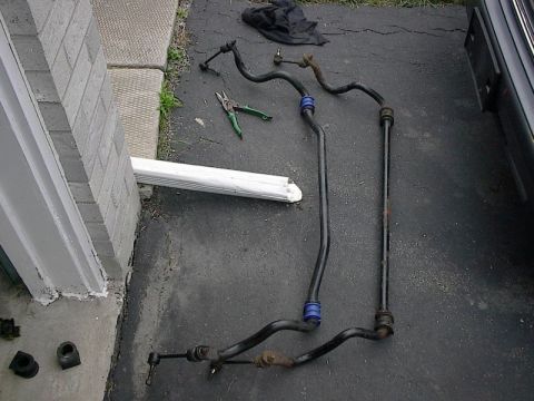 1985 Mercury Grand Marquis - Several car suspension components, including sway bars and end links, are arranged on an asphalt surface next to a brick structure.