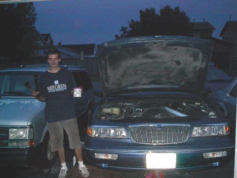 1985 Mercury Grand Marquis - A man stands with tools next to a blue sedan with its hood open, with another vehicle visible to the left.