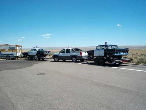 2001 Ford Crown Victoria - A 1956 police car is transported on a trailer, towed by a modern pickup truck, with another police car on a trailer in the background.