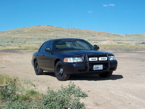 2001 Ford Crown Victoria - Black 2001 Ford Crown Victoria police car with push bar and PIAA lights parked on a dirt road under a clear blue sky.