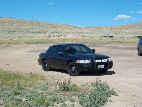 2001 Ford Crown Victoria - A dark sedan equipped with a push bar and auxiliary lights is parked in a dusty, outdoor lot.
