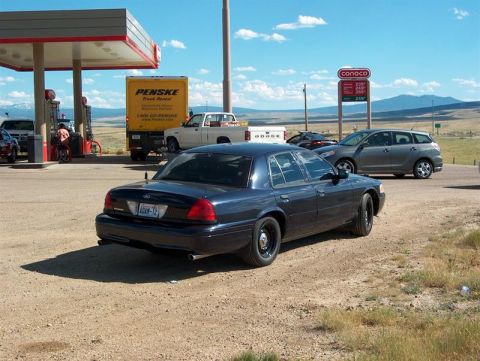 2001 Ford Crown Victoria - A dark 2001 Ford Crown Victoria sedan is seen from the rear at a gas station, with a Penske truck and other vehicles in the background.