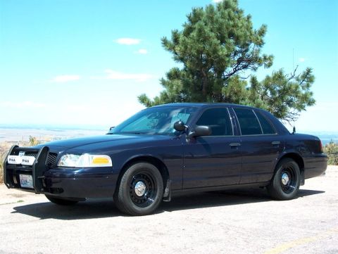 2001 Ford Crown Victoria - A dark blue 2001 Ford Crown Victoria police car is parked on a dirt lot with a tree and distant landscape in the background.