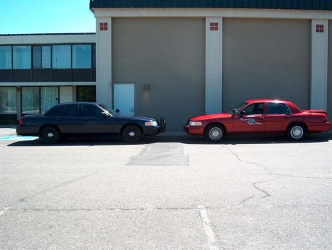 2001 Ford Crown Victoria - Two Ford Crown Victoria police cars, one black and one red, are parked side-by-side in front of a building.