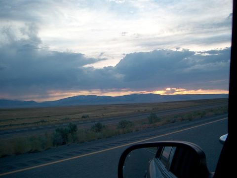 2001 Ford Crown Victoria - View from a vehicle's side mirror shows a highway stretching towards distant mountains under a dramatic sunset sky.