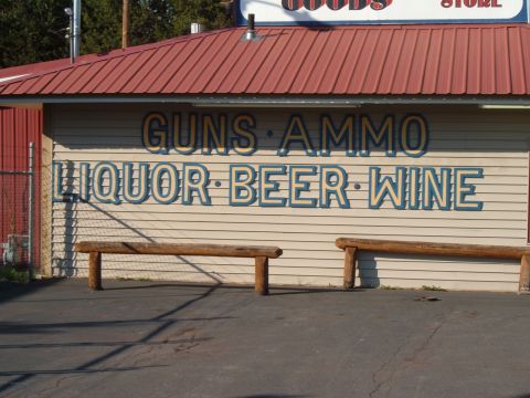 1989 Ford Bronco - A storefront displays prominent signage for various goods including guns, ammo, liquor, beer, and wine, with wooden benches outside.
