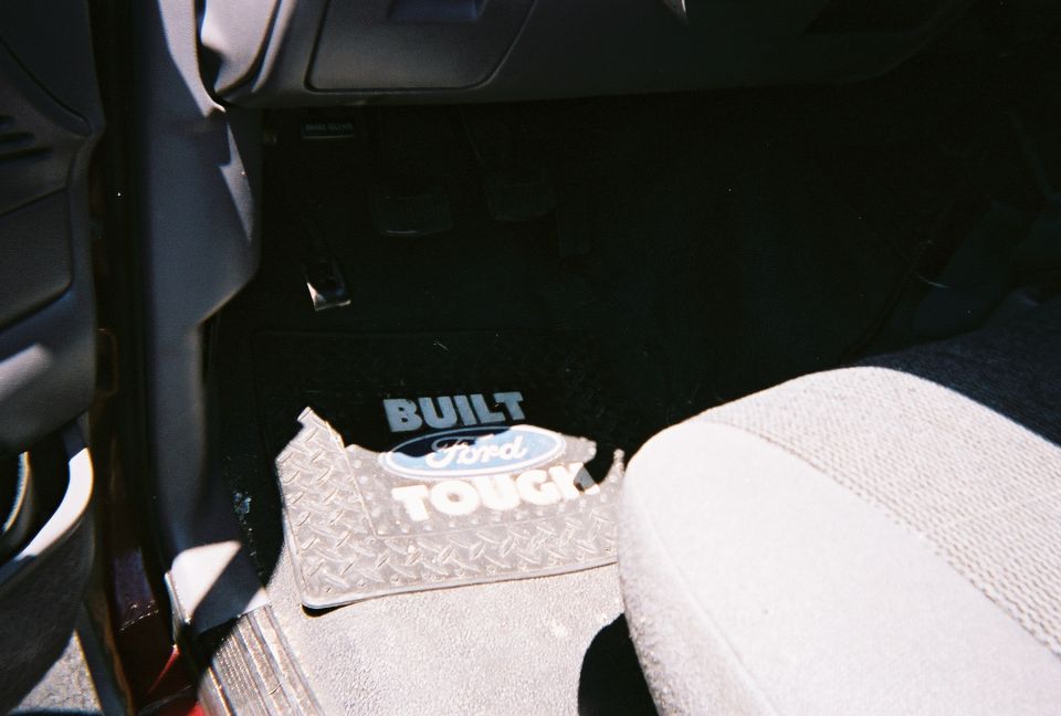 Driver floorboard with black Built Ford Tough rubber floor mat and gray cloth seat edge