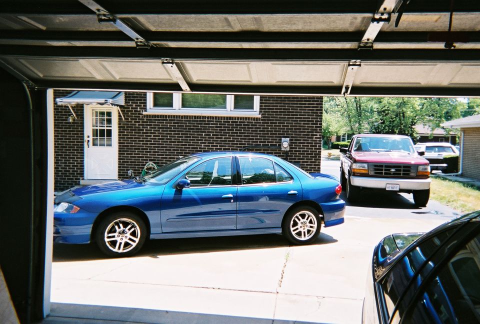 Blue Chevrolet Cavalier sedan parked in a driveway with a red pickup behind, viewed from a garage