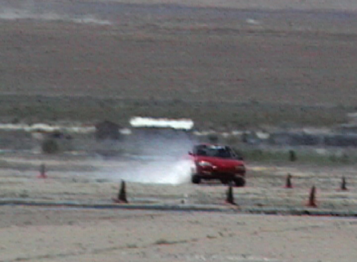 Red car drifting on a dirt track, kicking up smoke. Cones mark the course. Low resolution