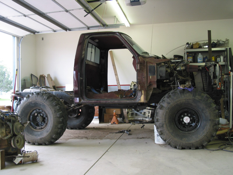 Maroon truck cab on bare frame with large mud tires mid-build in a garage