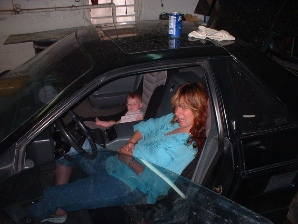 Black 1984 Pontiac Fiero interior with a woman in the driver's seat and a baby in the passenger seat
